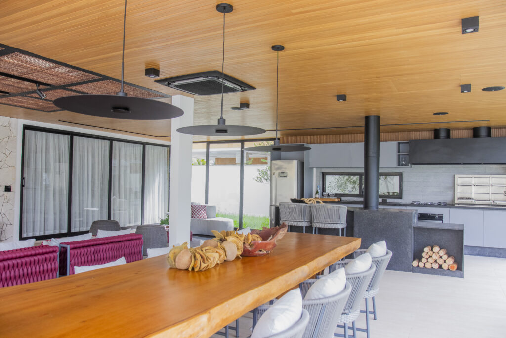 Área gourmet integrada com móveis sob medida Casttini, teto amadeirado, luminárias pendentes modernas e mesa de jantar em madeira de demolição, unindo design contemporâneo e elementos naturais.