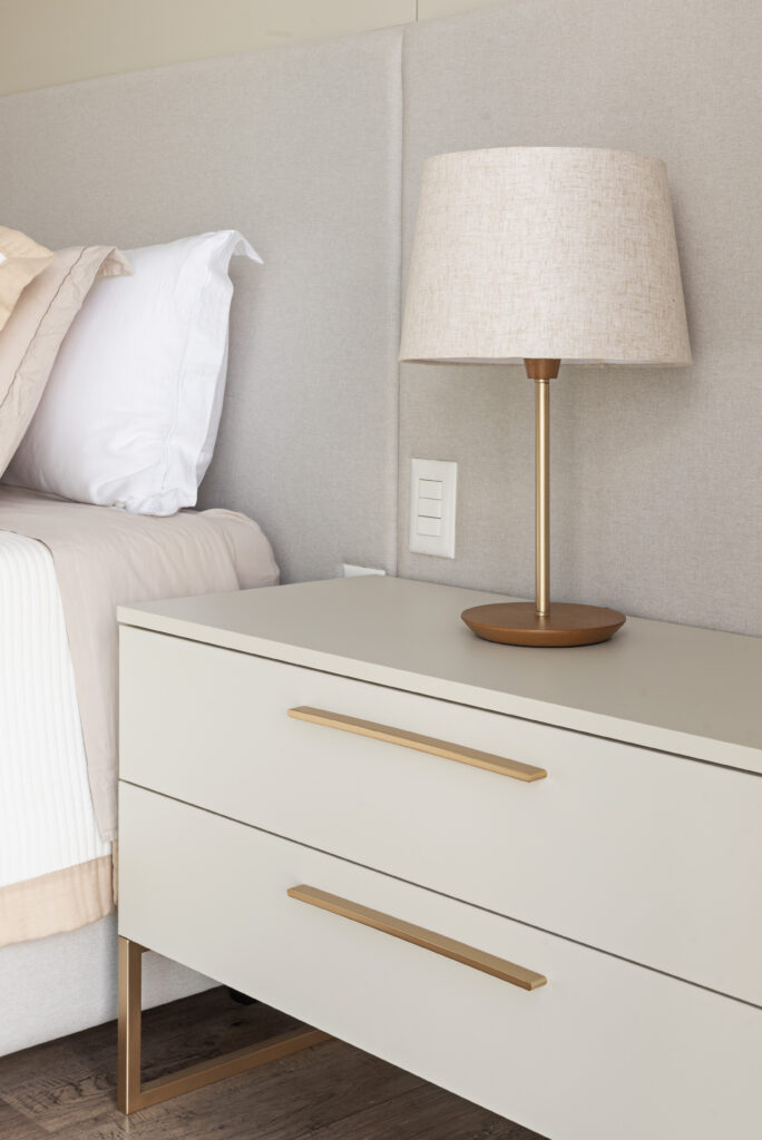 Mesa de cabeceira off-white com puxadores dourados em dormitório planejado Casttini, apresentando luminária sobre o móvel e painel estofado ao fundo para um ambiente acolhedor.