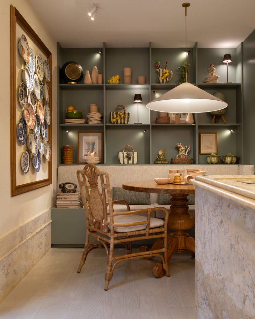 Cozinha planejada Casttini com mesa redonda de madeira, cadeira de vime e banco estofado integrado. Ao fundo, marcenaria sob medida em tom verde menta com prateleiras iluminadas exibindo louças decorativas e quadros, criando uma atmosfera mediterrânea e acolhedora.