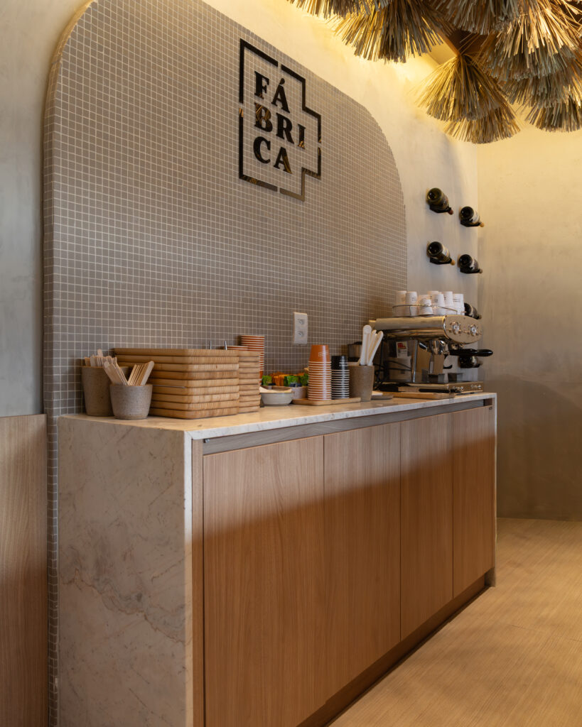 Detalhe do balcão de café e estação de espresso no Café Fábrica, evidenciando o acabamento Rovere da marcenaria sob medida Casttini, com texturas naturais de madeira, tampo de mármore e parede revestida em pastilhas cinzas com o logotipo da marca.