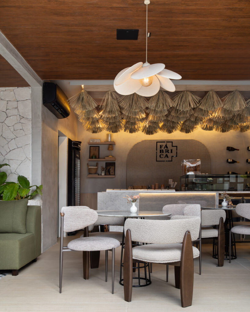 Ambiente interno do Café Fábrica projetado por Augusto Franco, destacando mesas de mármore com cadeiras estofadas, sofá verde, iluminação aconchegante e teto decorado com palha natural, apresentando os móveis planejados e a marcenaria sob medida da Casttini.