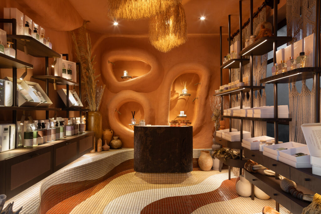 Ambiente Terra ao Lar na CASACOR Goias 2026 para a loja Cravo e Canela Aromas. O espaco exibe prateleiras iluminadas com aromatizadores de ambientes e cosmeticos, balcao central texturizado escuro, paredes de argila terra cota com nichos organicos esculpidos e piso de pastilhas onduladas.