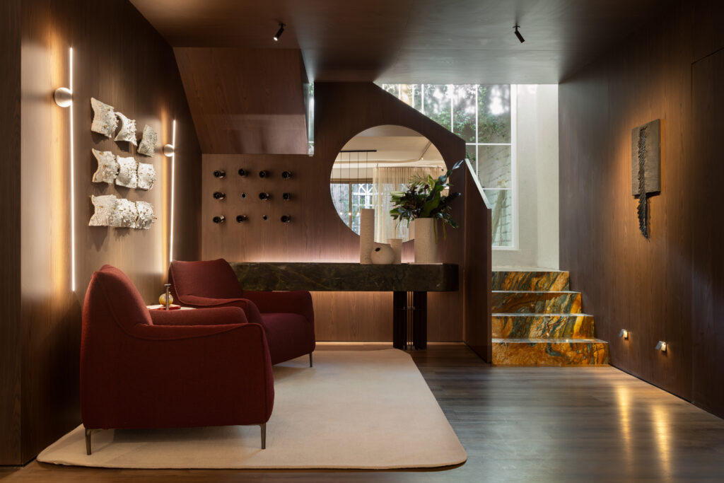 Sala Decantar por Leo Magno com marcenaria sob medida Casttini em acabamento Palissandro, poltronas confortáveis em tom vinho, tapete claro e estante com prateleiras iluminadas ao fundo.