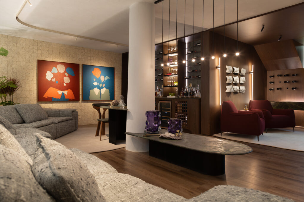 Vista ampla da Sala Decantar com mobiliário planejado Casttini, sofá modular cinza em destaque na frente, painéis com obras de arte coloridas na parede e estante de vinhos iluminada ao fundo.