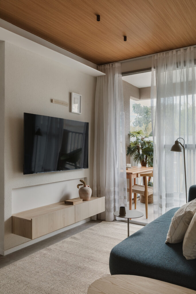 Sala de estar com rack de tv suspenso de madeira clara, cortinas brancas, tapete neutro e sofa azul em destaque, evidenciando a continuidade do piso e a leveza do ambiente por Casttini Moveis Planejados.
