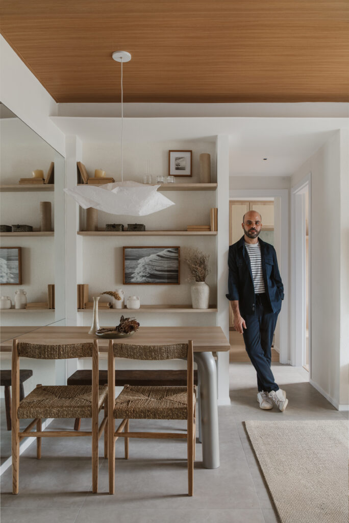 Homem em pé ao lado da mesa de jantar em ambiente integrado com cadeiras de palha, prateleiras decorativas e teto amadeirado em projeto da Casttini Moveis Planejados.