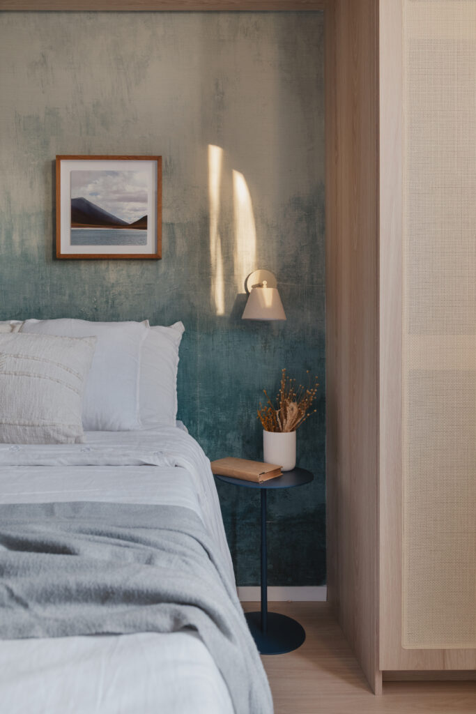 Dormitorio planejado com cabeceira de madeira natural, abajur aceso, parede verde com efeito de textura e painel de palha palha em projeto da Casttini Moveis Planejados.