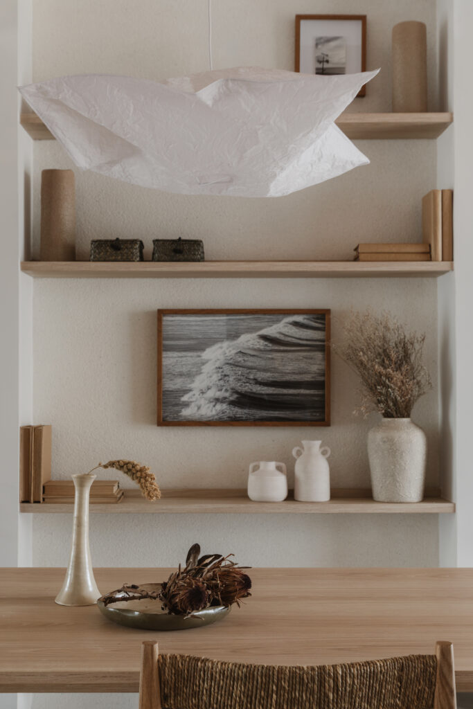 Mesa de madeira com vaso e luminaria pendente de papel em ambiente decorado com prateleiras e quadro com foto de onda, destacando acabamento continuo e fluidez visual Casttini Moveis Planejados.